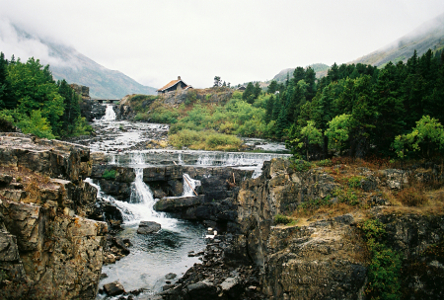 [This is a multi-level waterfall. At the top is a narrow section of falls which then widens as it runs across a near-level section of land. A second falls section is in several parts as all the water runs across a short drop then it funnels through three holes in the rock as it drops the next section to a pool of water.]